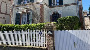A two-story building with a white facade and red brick accents, featuring multiple windows with shutters, a stone entrance, and a white picket fence.