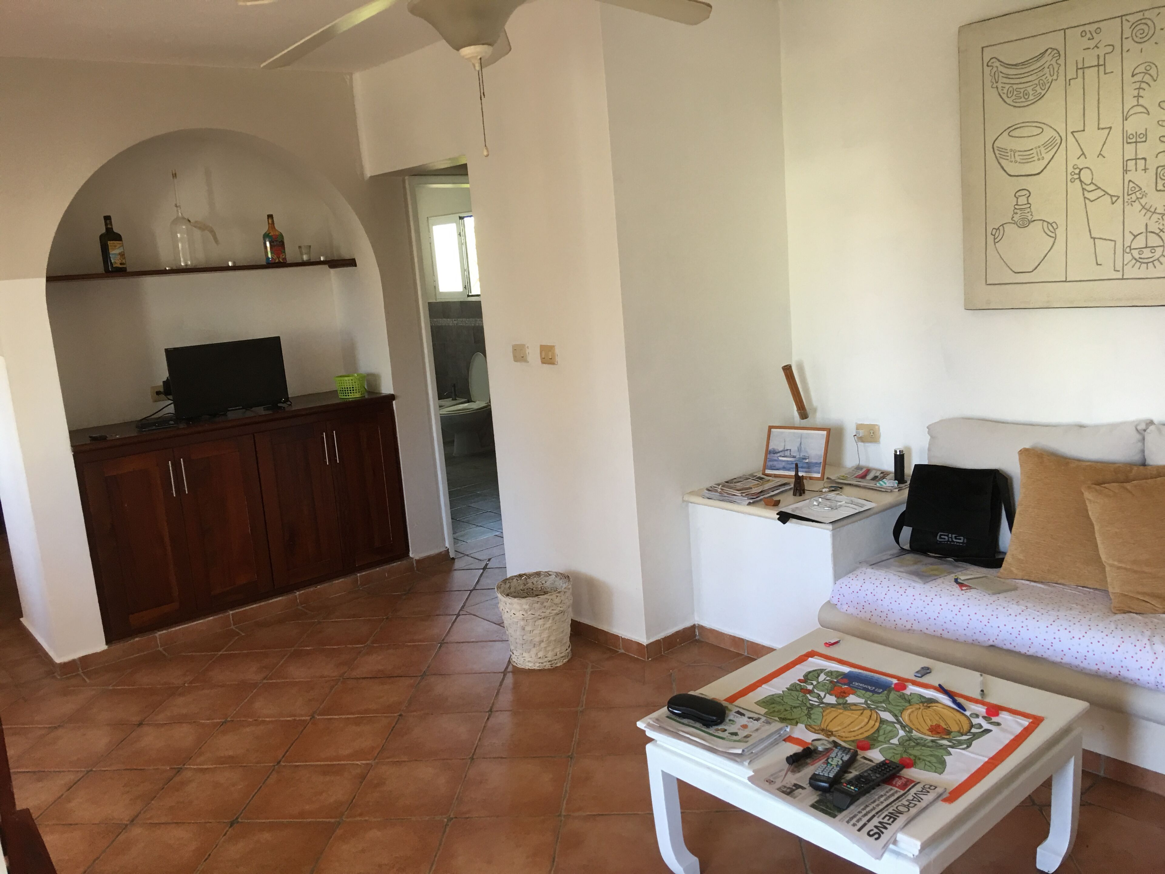 Una sala de estar con un sofá blanco, una mesa de centro con periódicos y controles remotos, un televisor sobre un mueble de madera y un cuadro en un pequeño atril.