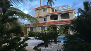 Un bâtiment à deux étages, doté d’un toit en tuiles, entouré de palmiers et bordé d’une piscine.