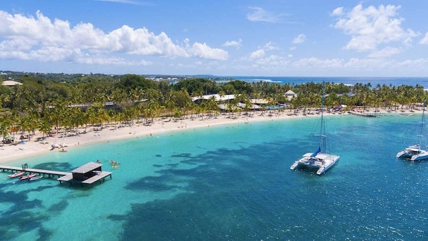 Vue aérienne d’un complexe hôtelier en bord de mer, avec une eau turquoise et limpide, une plage de sable et plusieurs bateaux ancrés près du rivage.