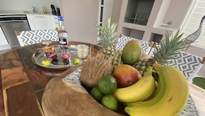Une table en bois sur laquelle se trouve un panier de fruits contenant des bananes, des citrons verts, une mangue et un ananas. On peut également apercevoir une bouteille de rhum jamaïcain et un pot de miel.
