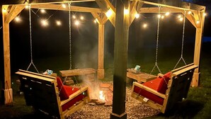En træpavillon med to gynger, lyskæder og et bålsted.