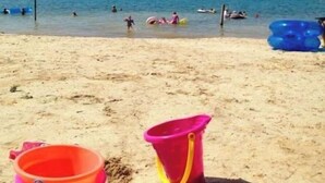 En strand med klart, blåt vand, folk der svømmer og leger, og et bjerglandskab i baggrunden.