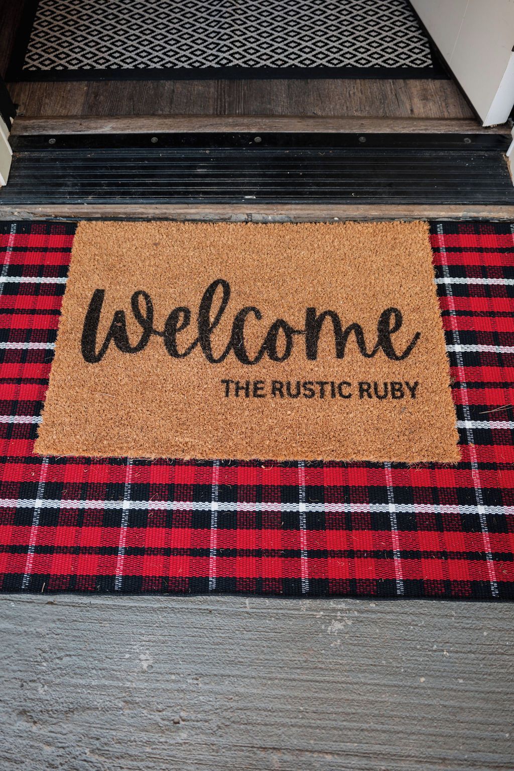 A welcome mat with "Welcome The Rustic Ruby" text on a red and black checkered doormat.