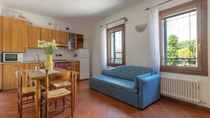 A compact living space with a kitchenette, a dining area with wooden chairs, a blue sofa, and large windows with curtains. - Apartment for 2 people surrounded by greenery! (Mira)