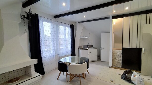 A modern kitchen with white cabinets, a stove, and a sink. There is a dining area with a round table and chairs. - Fully Equipped Furnished Design (Poissy)