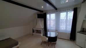 A compact living space with a kitchenette, a dining table with two chairs, a sofa, and a large window with curtains. - Fully Equipped Furnished Design (Poissy)
