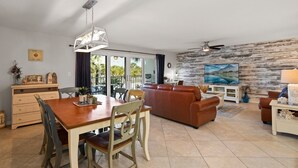 A spacious living area with a wooden dining table, a brown leather sofa, and a TV mounted on a textured wall.