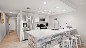 A modern kitchen with stainless steel appliances, a marble island, and white bar stools.