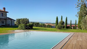 A swimming pool with a wooden deck, stone house, and cypress trees in the background. - Ribusieri Country House (Cinigiano)