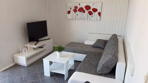 Ein modernes Wohnzimmer mit einem Flachbildfernseher auf einem weißen Schrank, einem grauen Sofa mit grauem Kissen, einem weißen Couchtisch mit Pflanze und einem weißen Beistelltisch.
