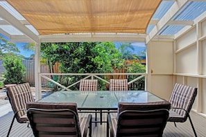 An outdoor patio with a glass table and striped chairs, surrounded by a wooden deck and a covered area with a view of greenery.