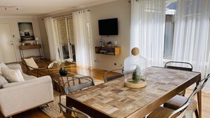 Ein geräumiges Wohnzimmer mit Esstisch aus Holz, bequemen Sitzmöglichkeiten, einem Flachbildfernseher und einer Glastür, die auf einen Balkon führt.