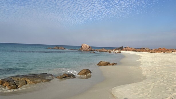 Ein Strand mit klarem, blauem Wasser, weißem Sand und vereinzelten Felsen.