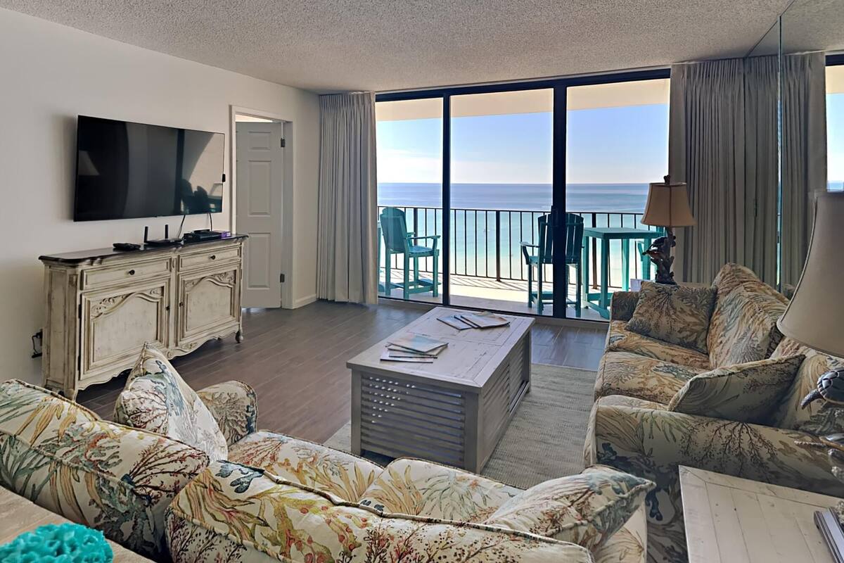 A living room with a large TV, a sofa, a coffee table, and a balcony with a view of the ocean.