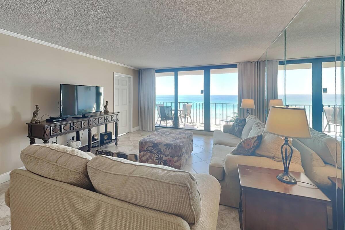 A spacious living room with a large flat-screen TV, comfortable seating, and a balcony with a view of the ocean.
