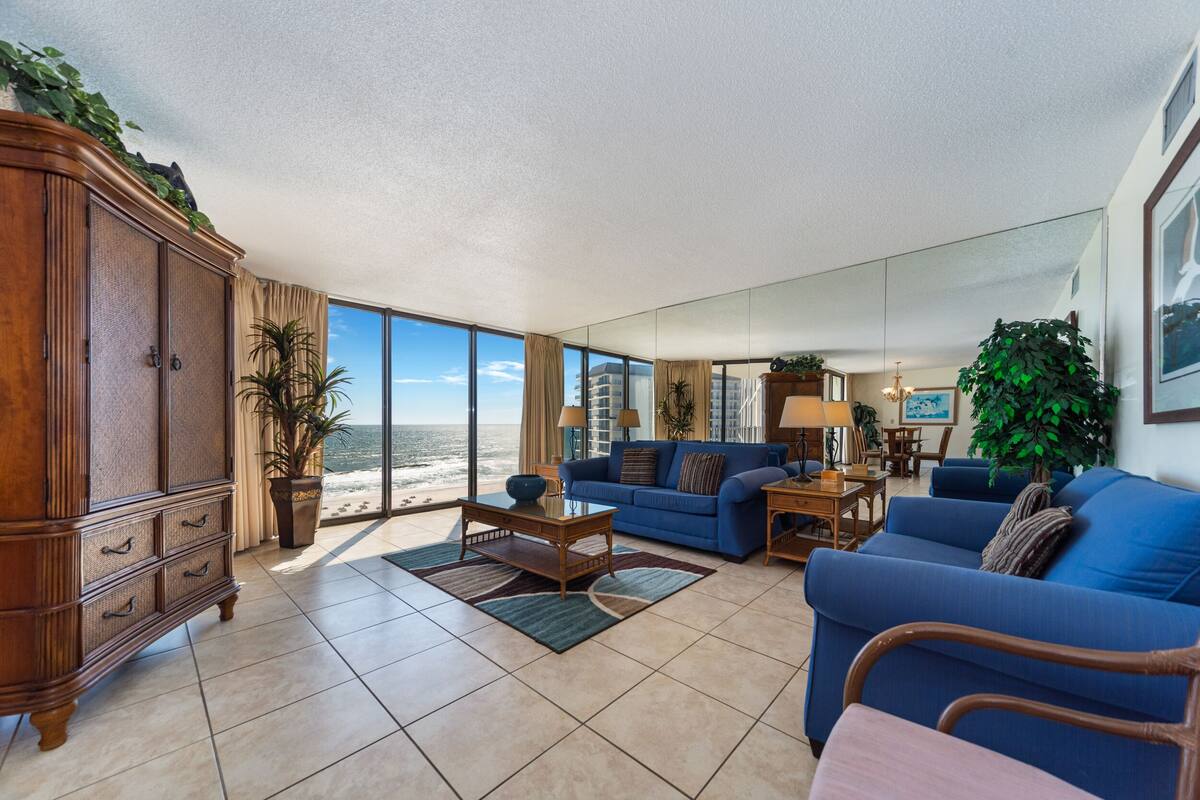 A spacious living room with a large window overlooking the beach, a blue sofa, a wooden cabinet, and a dining area with a table and chairs.