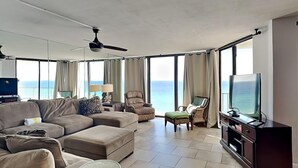 Un salón amplio con un sofá grande, una mesita redonda y una televisión. La habitación tiene ventanas grandes con cortinas, un ventilador de techo y vista al océano.