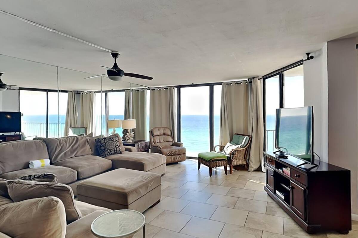 A spacious living room with a large sofa, a small round table, and a television. The room has large windows with curtains, a ceiling fan, and a view of the ocean.