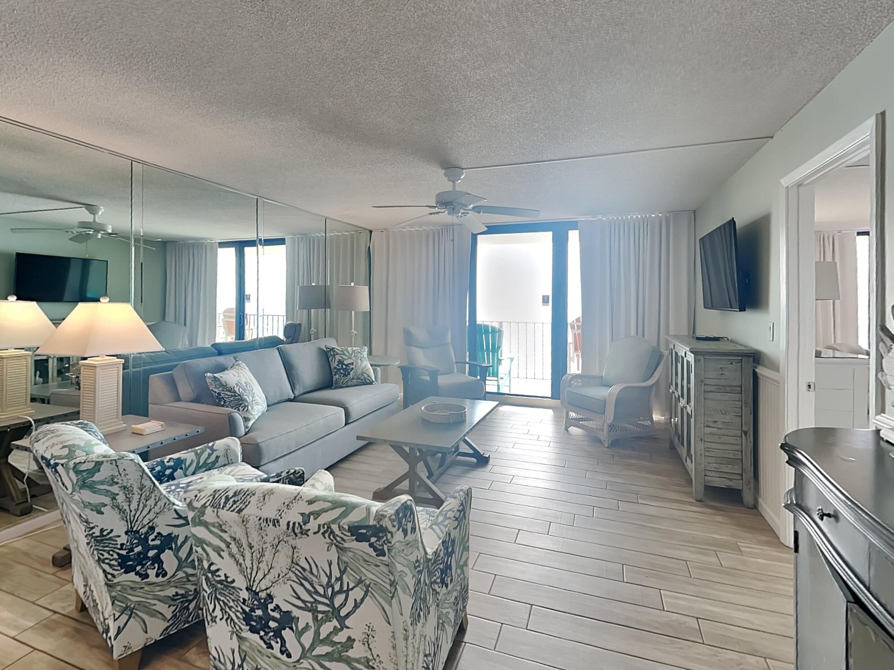 A modern living room with a sofa, armchairs, a coffee table, and a TV. There is a balcony with a view, and the room has a ceiling fan.