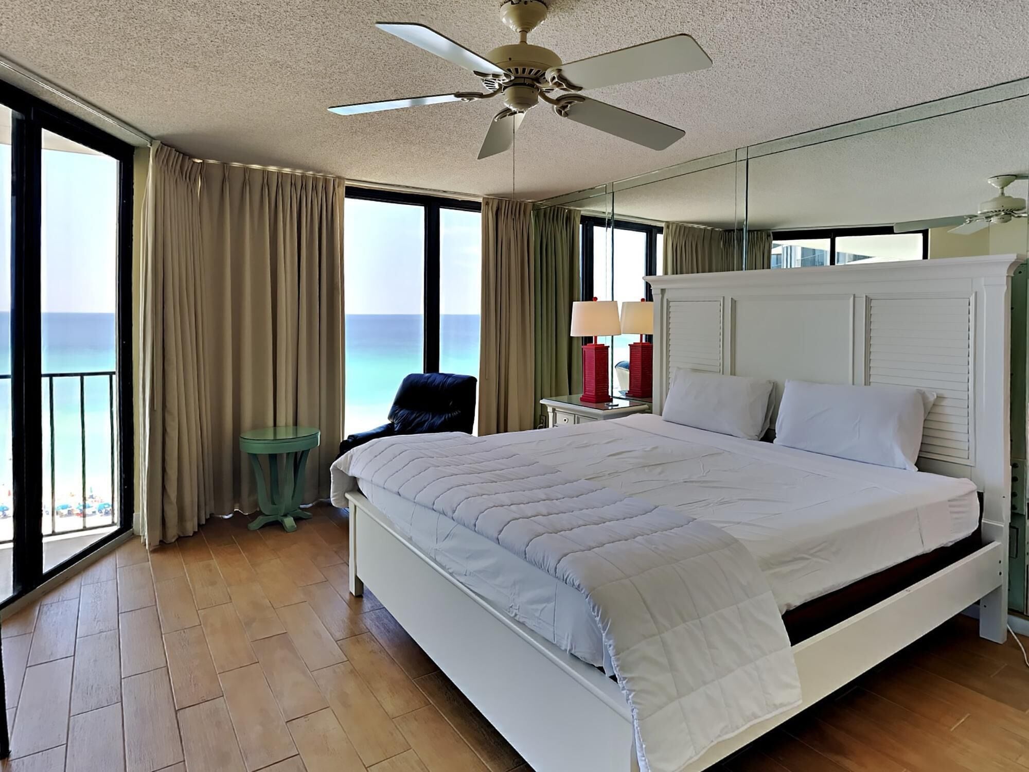 A hotel room with a large bed, a ceiling fan, a nightstand with lamps, a chair, and a view of the ocean.