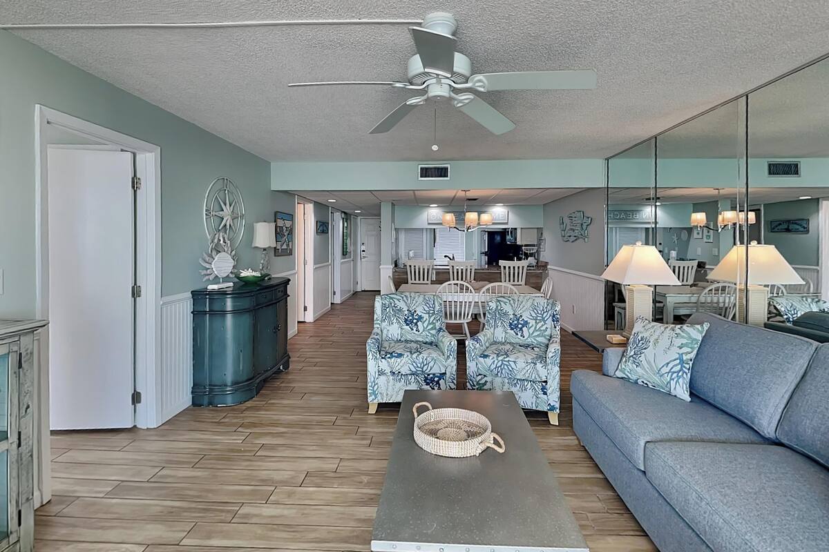 A spacious living room with a ceiling fan, a dining area with white chairs, and a kitchen in the background.