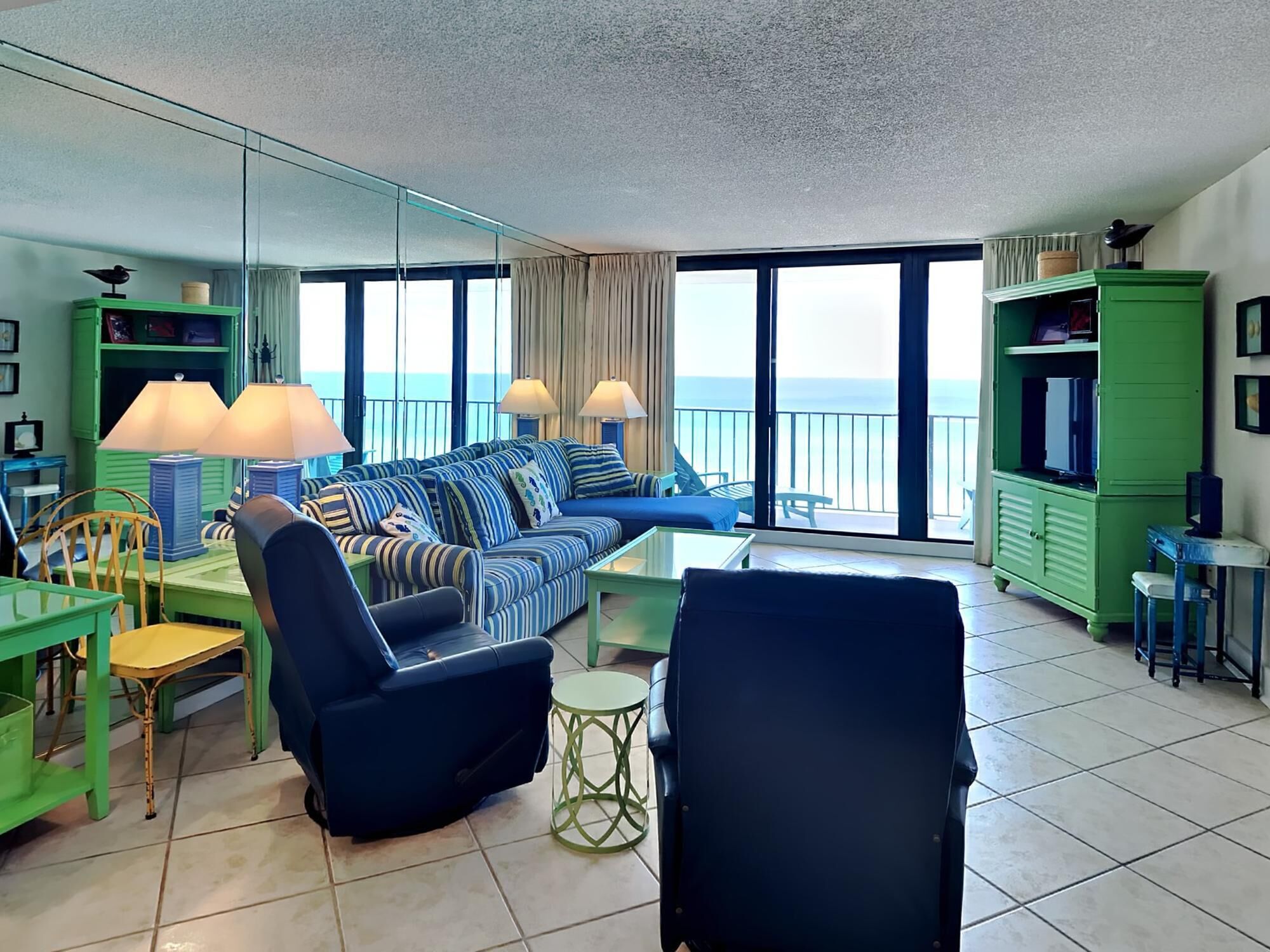 A modern living room with a large sofa, chairs, and a dining area. It features large windows with a view of the sea.