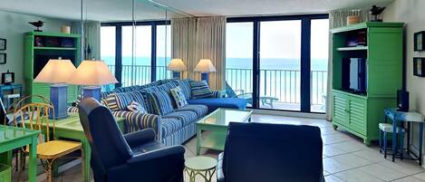 A modern living room with a large sofa, chairs, and a dining area. It features large windows with a view of the sea.