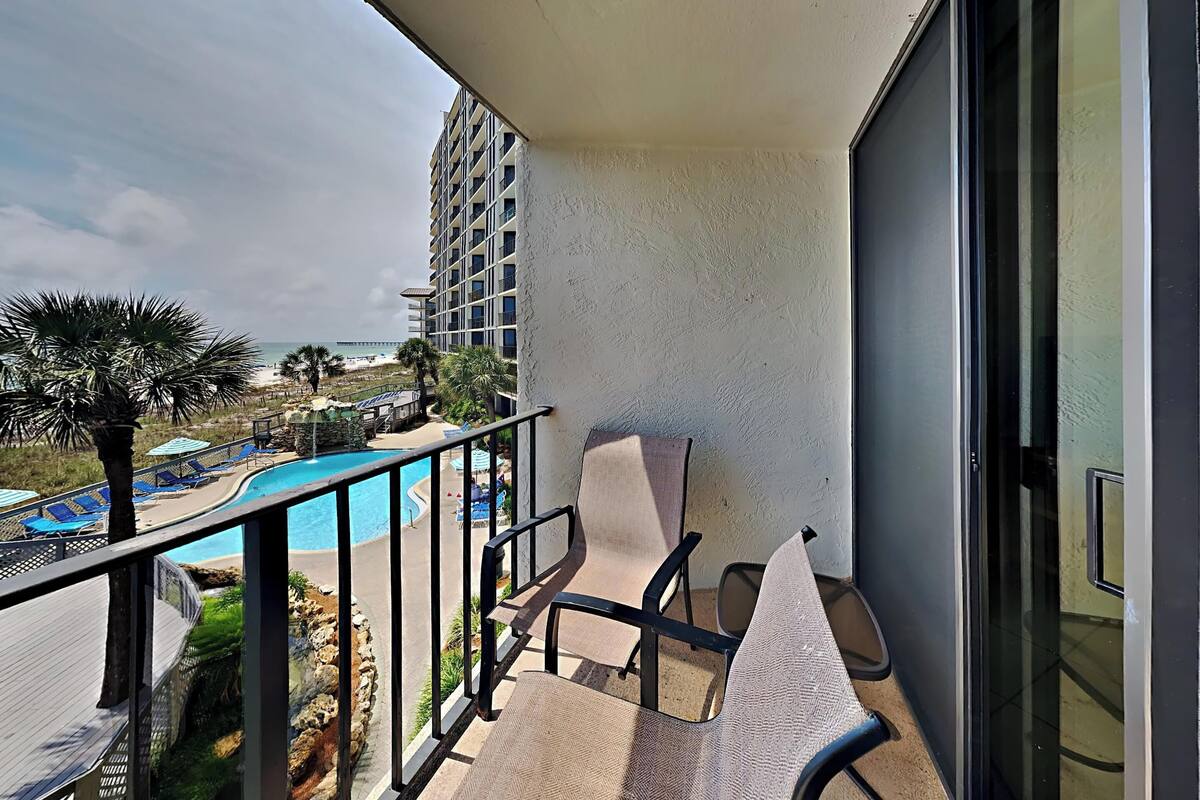 A balcony with a pool, lounge chairs, and a view of the beach and ocean.