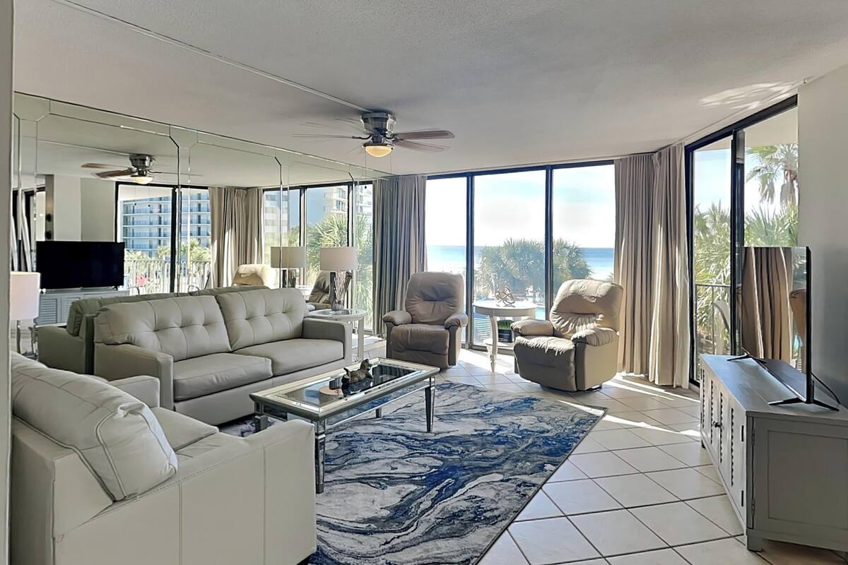 A spacious living room with a large window offering a view of the ocean, a ceiling fan, and a flat-screen TV.