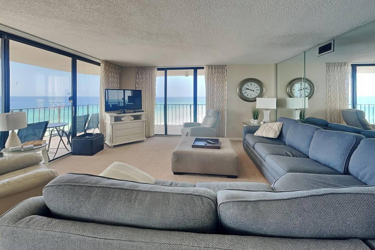 A spacious living room with a large sofa, a television, and a balcony with a view of the sea.