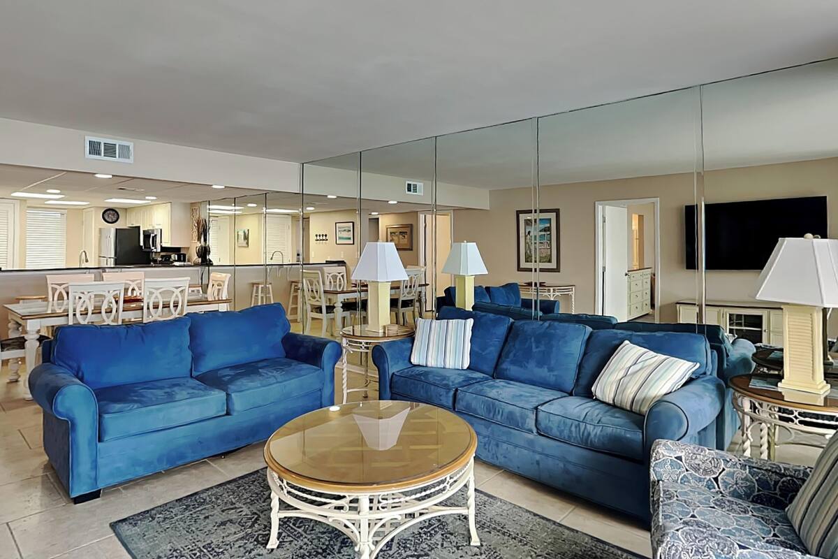 A living room with two blue sofas, a round coffee table, and a patterned armchair. A large mirror reflects the room, and a TV is mounted on the wall.