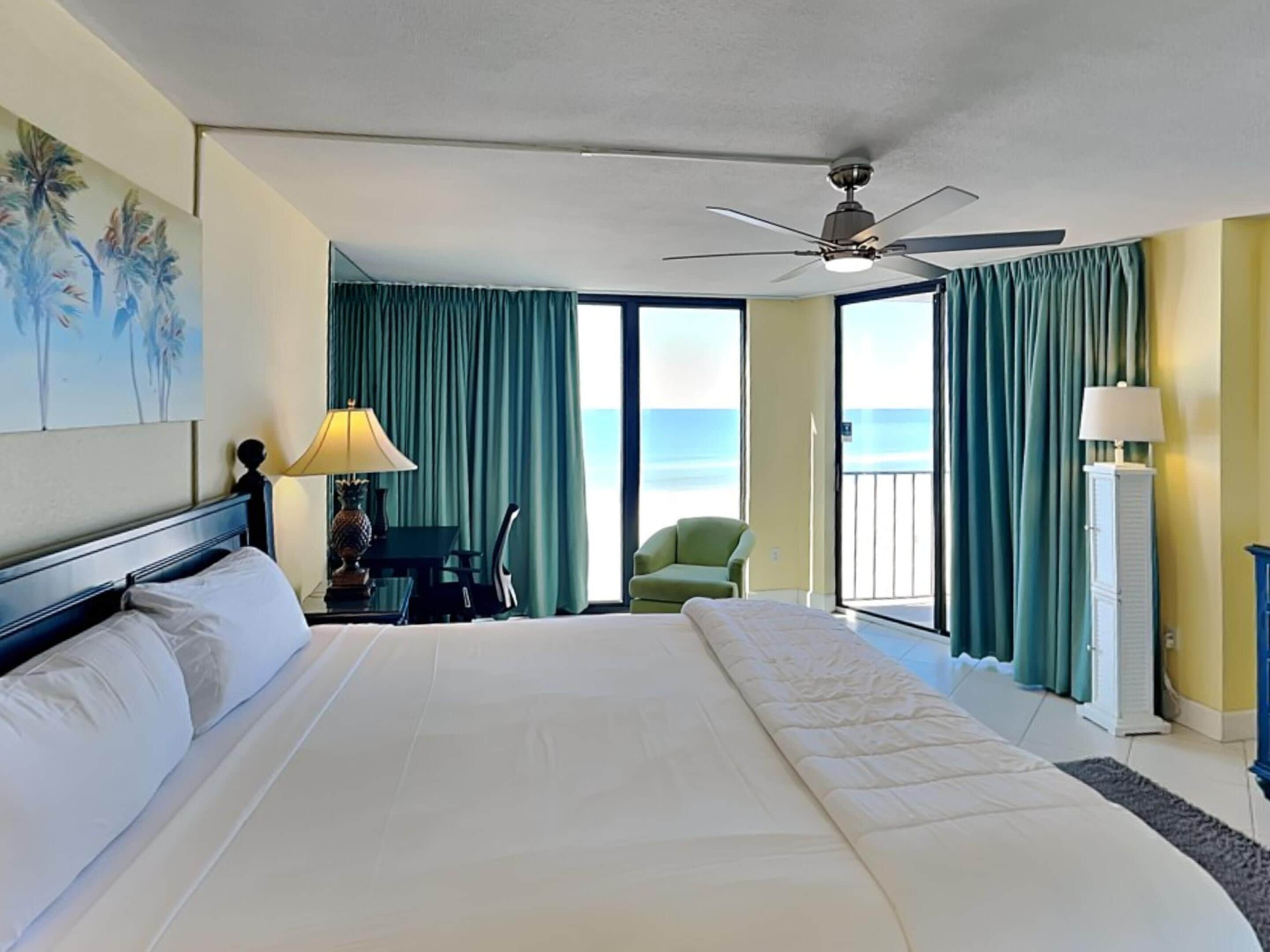 A hotel room with a large bed, a ceiling fan, a desk with a lamp, a chair, and a view of the ocean through the windows.