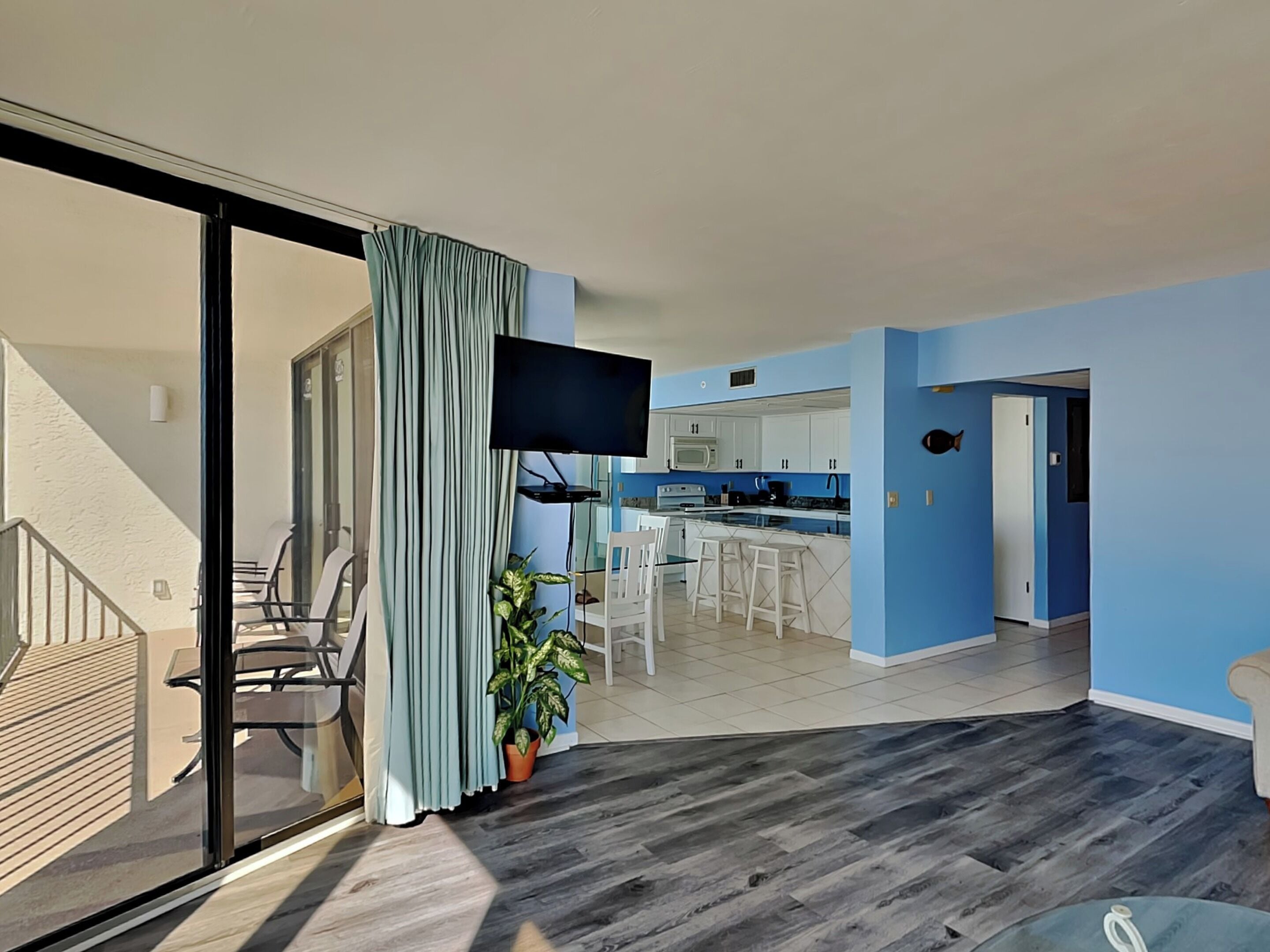 A modern living room with a large sliding glass door leading to a balcony, a kitchen with blue walls, and a flat-screen TV mounted on the wall.
