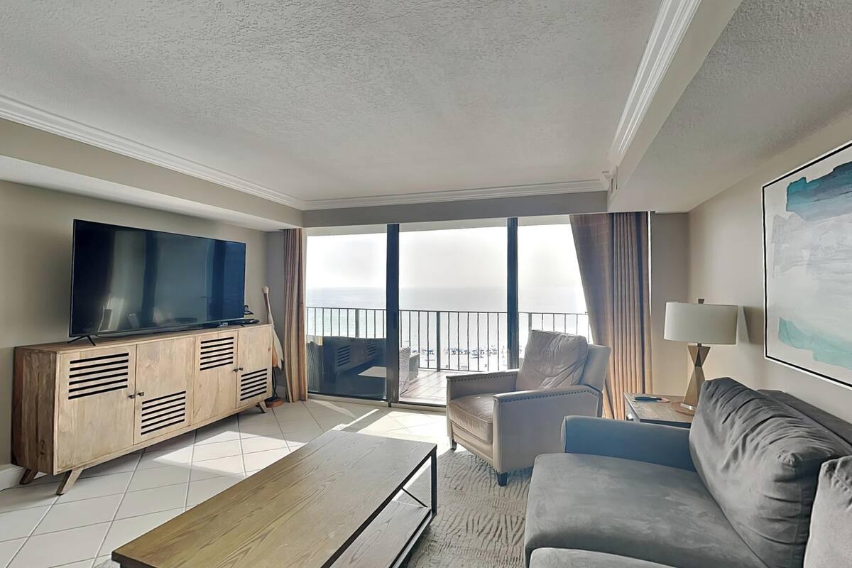 A modern living room with a flat-screen TV, a wooden cabinet, a sofa, a coffee table, and a balcony with a view.