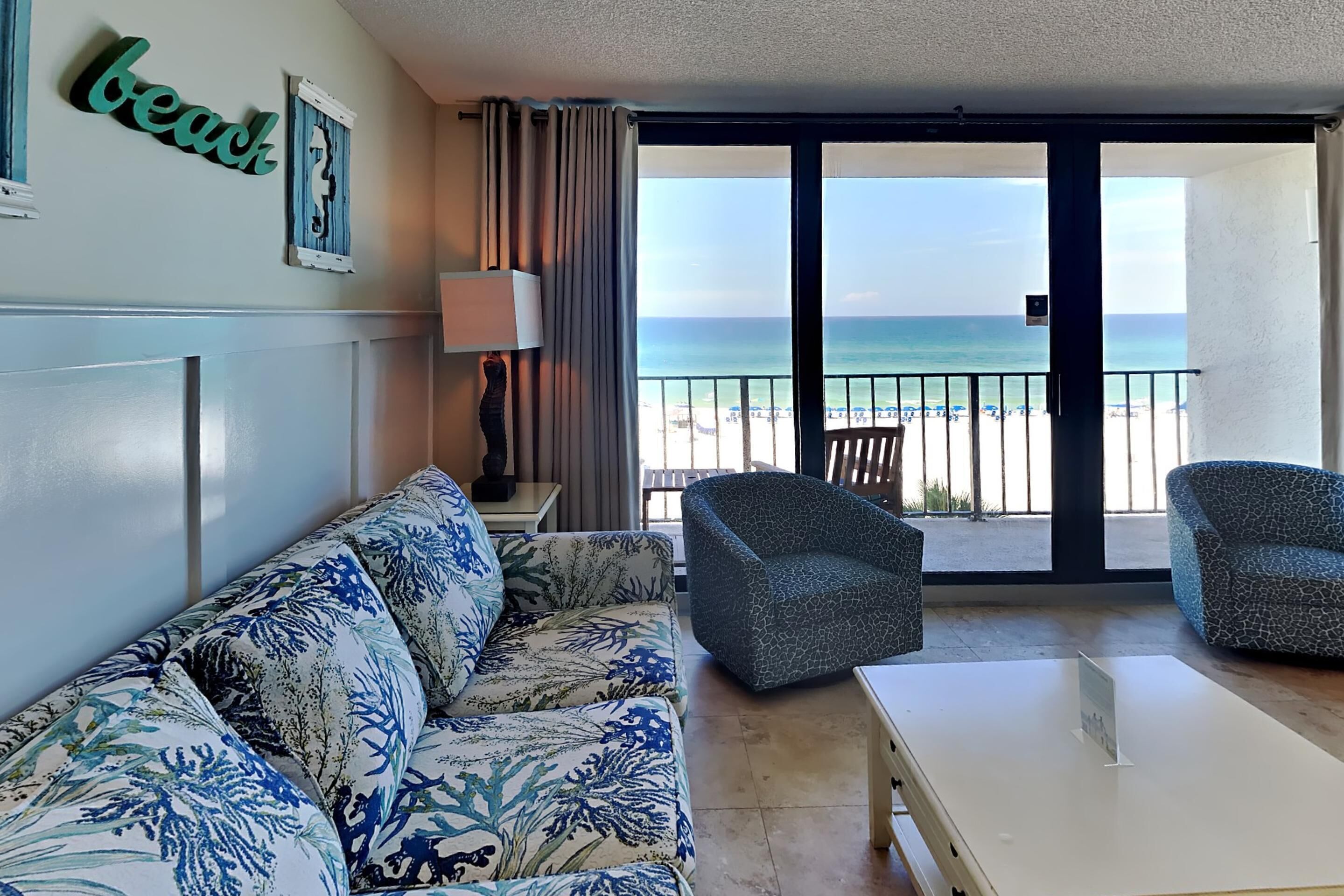 A room with a sofa, two armchairs, a table, a lamp, and a view of the beach.