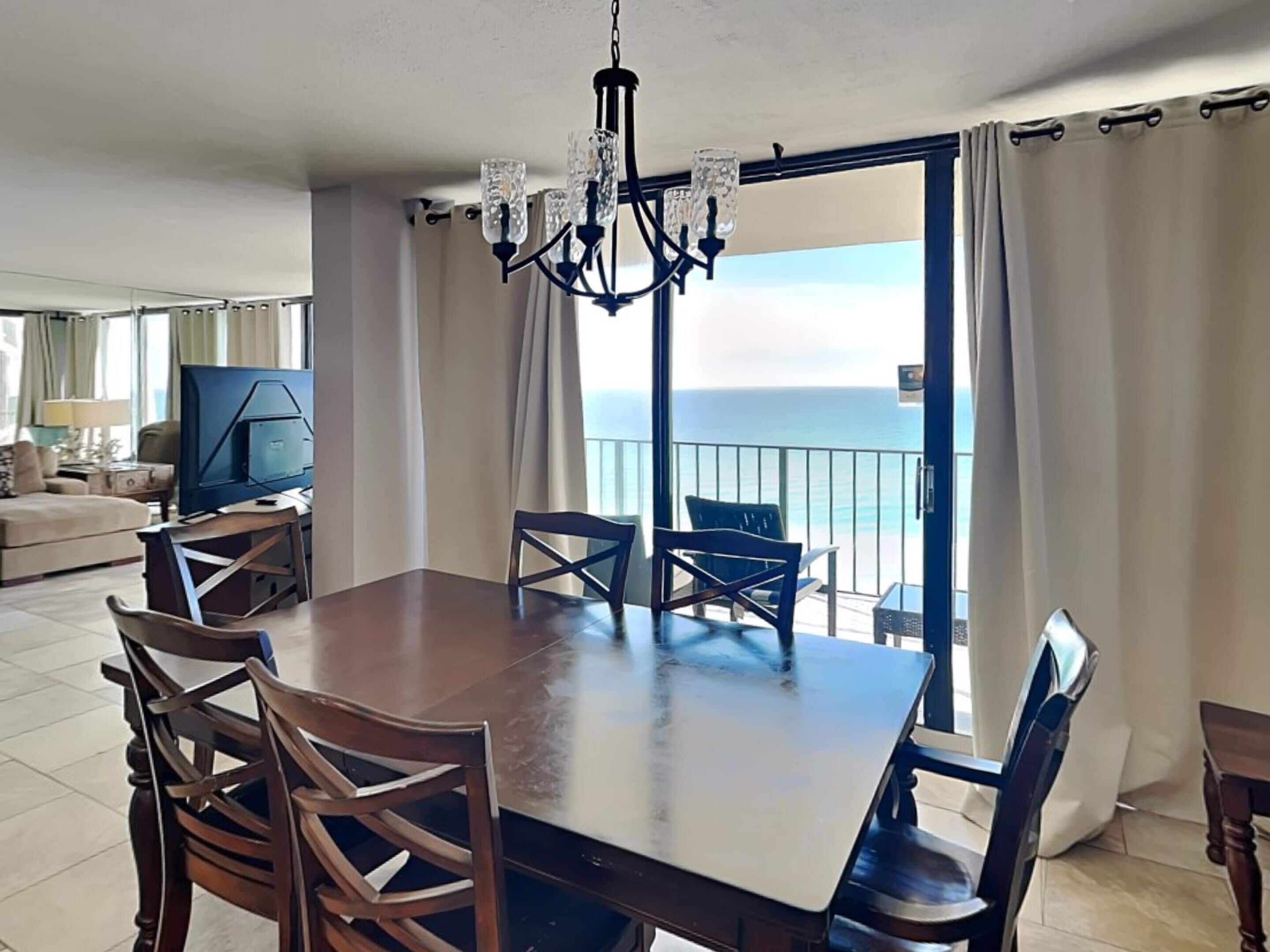 A dining area with a large table and chairs, a chandelier, and a view of the ocean through a balcony.