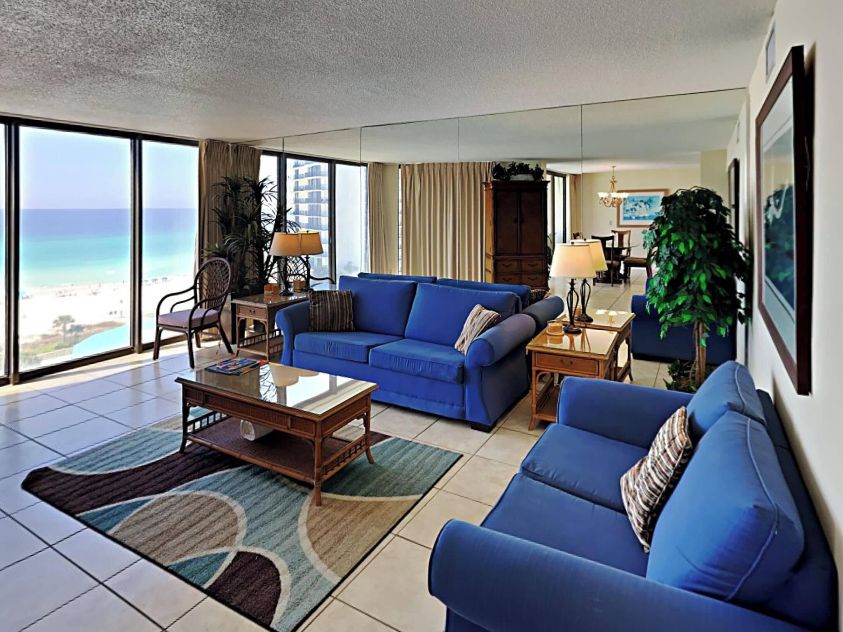 Ein geräumiges Wohnzimmer mit einem großen blauen Sofa, einem Couchtisch und Blick auf den Strand durch große Fenster.