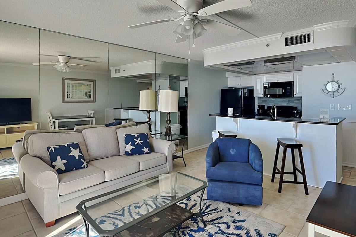A modern living room with a sofa, armchair, and coffee table. A kitchen area with appliances and a dining table is visible in the background.