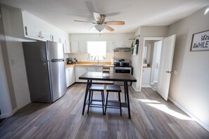 A modern kitchen with white cabinets, a stainless steel refrigerator, a wooden dining table with two stools, and a ceiling fan.