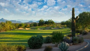Un campo de golf con búnkeres de arena, cactus y montañas al fondo.