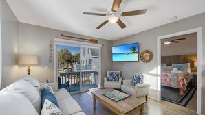 A living room with a sofa, coffee table, and a TV. There is a balcony with a view of palm trees and a pool. - AMI Moana Villa 3-minute walk to the beach plus heated pool, grill, beach gear (Holmes Beach)