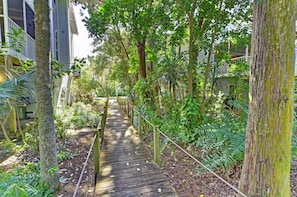 A wooden pathway surrounded by lush greenery and trees, leading to a building with multiple windows and balconies.
