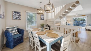 A dining area with a white table set with woven placemats, a blue wicker chair, and a hanging light fixture. - AMI Moana Villa 3-minute walk to the beach plus heated pool, grill, beach gear (Holmes Beach)