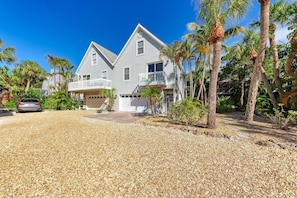 A two-story house with a balcony, surrounded by palm trees and a gravel driveway.