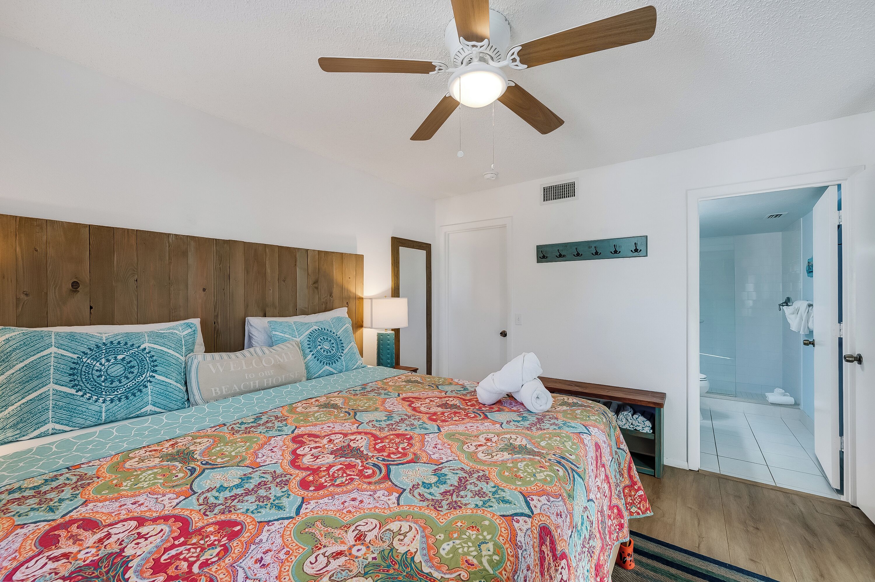 A bedroom with a large bed, a ceiling fan, a nightstand, a lamp, and a door leading to a bathroom.