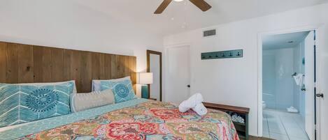 A bedroom with a large bed, a ceiling fan, a nightstand, a lamp, and a door leading to a bathroom.