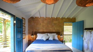 Un dormitorio con una cama grande, cabecera de madera y pared de ladrillo. Hay una puerta azul y una ventana con vistas a la vegetación.