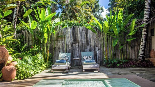Zona de piscina con dos sillones de descanso, valla de madera y plantas tropicales.