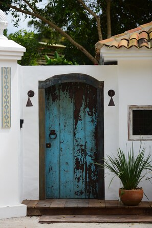 Une porte bleue, usée par les intempéries, dotée d’une poignée en métal, flanquée de deux appliques murales et agrémentée d’une plante en pot à côté.